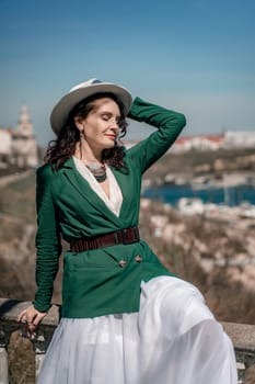 Woman walks around the city, lifestyle. A young beautiful woman in a green jacket, white skirt and hat is sitting on a white fence with balusters overlooking the sea bay and the city