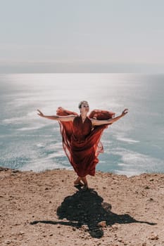 Woman red dress sea. Female dancer posing on a rocky outcrop high above the sea. Girl on the nature on blue sky background. Fashion photo