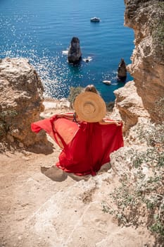 woman in a red dress stands on a cliff overlooking the ocean. The scene is serene and peaceful, with the woman's hat and dress adding a touch of elegance to the image