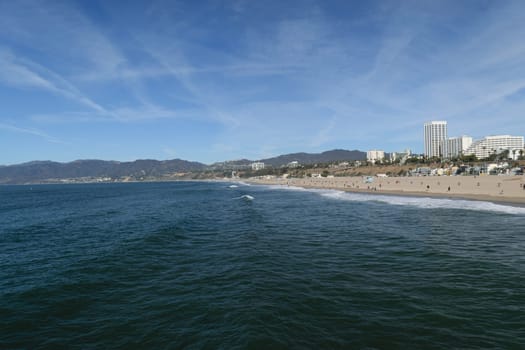 A scenic view of a beach with clear blue water and a cityscape in the background. The sky is clear with a few wispy clouds. People are walking along the sandy shore.