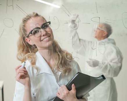 A woman chemist writes a formula on glass. An elderly Caucasian man in a protective suit is doing tests