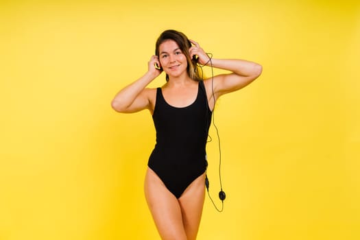 Caucasian Woman in Black Bodysuit Listening Music in Headphones On Yellow Background.