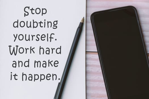 Motivational quote written on notebook with pen and smartphone on wooden background - Stop doubting yourself. Work hard and make it happen.