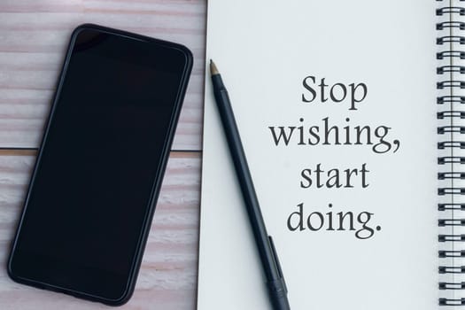 Motivational quote written on notebook with pen, and smartphone on wooden background - Stop wishing start doing.
