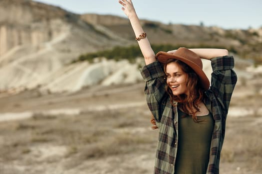 empowered woman in plaid shirt and hat raises arms triumphantly in the vast desert landscape