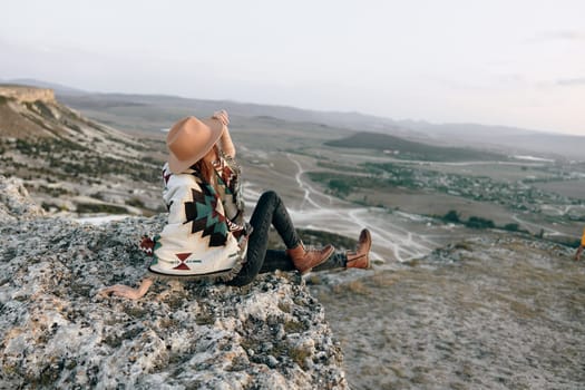 Serene woman with hat and backpack sits atop mountain summit, enjoying breathtaking view