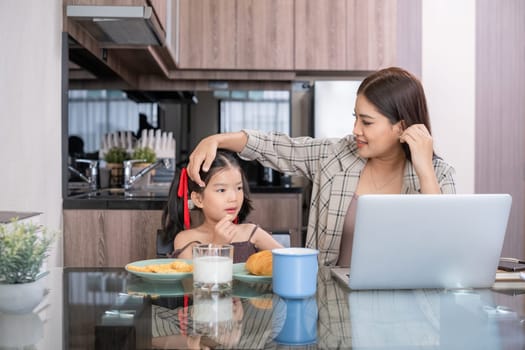 Asian mother working on laptop while interacting with daughter in kitchen. Concept of balancing work and family time.