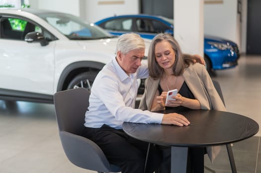 A mature couple is sitting in a car dealership and looking at a smartphone