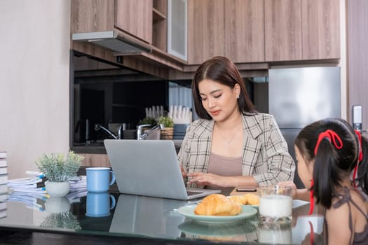 Asian mother working on laptop in kitchen with daughter nearby. Concept of work-life balance and remote work.