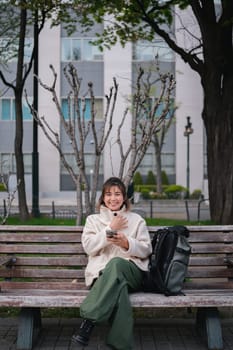 Woman sitting on a park bench smiling while using a smartphone with a backpack. Concept of outdoor relaxation and digital connectivity.
