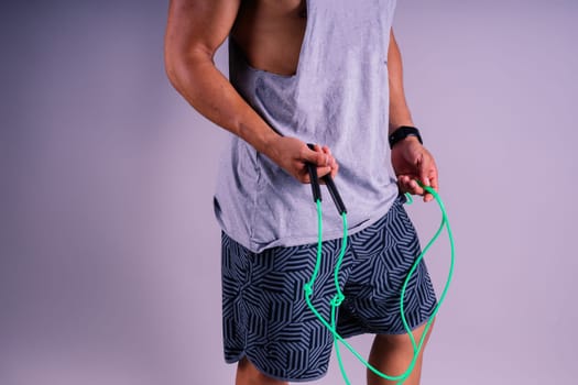 A man with a grey tank top is holding a green jump rope with his elbow bent and waist twisted, showing off his toned chest and wrist strength