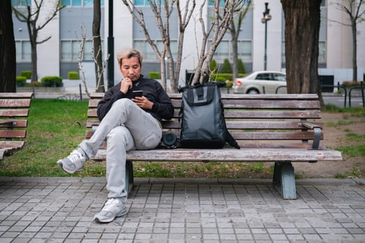Man sitting on a park bench using smartphone with backpack and camera. Concept of urban relaxation and technology.