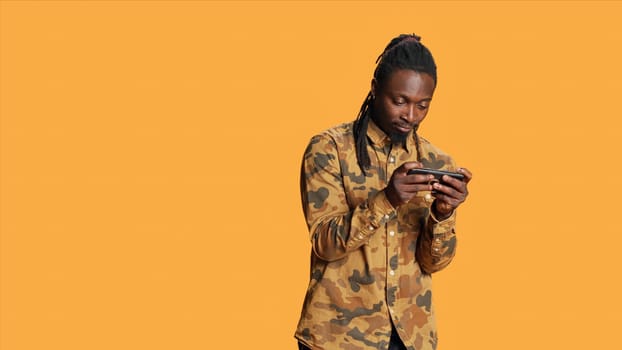 African american guy having fun with videogames online on mobile device, playing rpg contest standing over orange background. Cheerful gamer enjoying gaming tournament in studio.