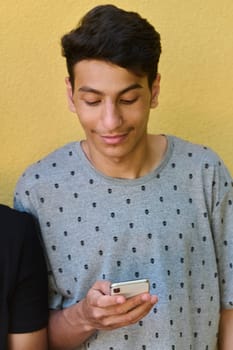 Middle Eastern teenager engrossed in his mobile phone while leaning against a vibrant yellow wall.