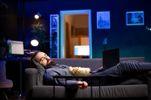 Man sleeping on couch from tiredness after working on laptop all day before managing to watch TV and enjoy bowl of popcorn. Freelancer falls asleep while solving tasks on notebook and looking at film