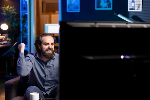 Ecstatic sports fan watching live match broadcast sitting on couch in apartment, celebrating favorite team win. Overjoyed man relishing about own country victory in world cup football competition