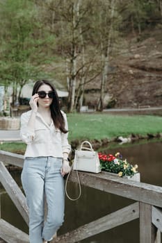 Portrait of one beautiful Caucasian brunette girl in sunglasses and stylish clothes stands on a wooden bridge with flowers against the backdrop of a blurred river and mountain in a nature reserve on a spring day in Belgium, close-up side view.