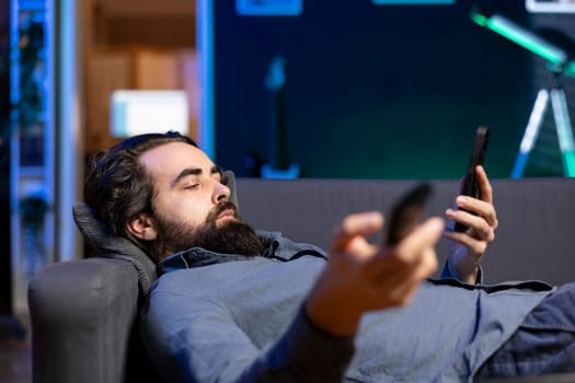 Homeowner bored at home laying down, using TV to watch movies on streaming services and chatting with friends. Apathetic man lounged on couch, mindlessly consuming entertainment and using mobile phone