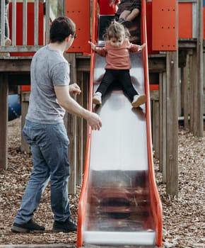 Portrait of one young Caucasian male father watching over his little daughter as she slides down a slide on a playground in a nature reserve on a sunny spring day in Belgium, close-up side view.