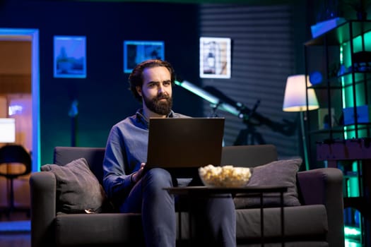 Man enjoying bowl of popcorn while watching cable programs on TV and sending emails on laptop. Spectator on couch in living room eats snacks during movie and works on notebook