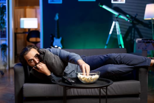 Person hugging pillow while binging suspenseful TV series on subscription based streaming services, enjoying popcorn. Scared man at home watching horror VOD shows on television display, eating snack