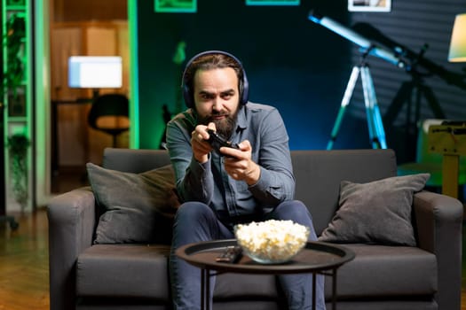 Man in neon lit apartment playing videogames on gaming console with teammates. Player fighting foes in esports game using joystick, discussing with friends through headset