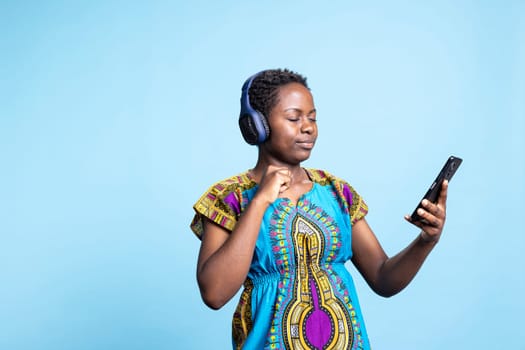 Female model playing music from a playing in the studio, having fun with groovy music on audio headset over blue background. Smiling confident woman listening to tunes on headset.