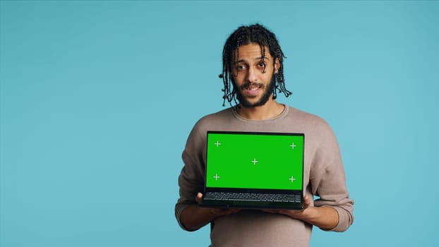 Joyous BIPOC man holding green screen laptop, having positive mood. Smiling person presenting chroma key notebook, doing recommendation, isolated over studio background, camera A