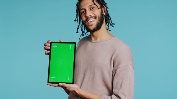 Joyful man holding green screen tablet, showing positive emotion. Upbeat person presenting chroma key device, doing recommendation, isolated over studio background, camera B close up