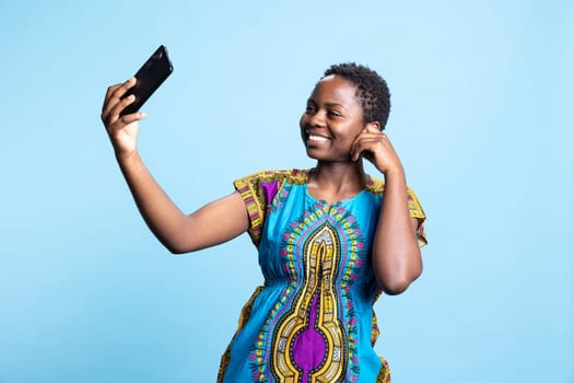 African american woman smiling at smartphone camera in studio, taking pictures and posting on social media apps. Female model photographing herself over blue background, ethnic style.