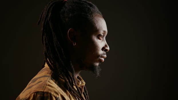 African american guy looking at camera with confidence, feeling happy and relaxed with dreads and cool clothing items. Model posing, standing over black background smiling. Handheld shot.