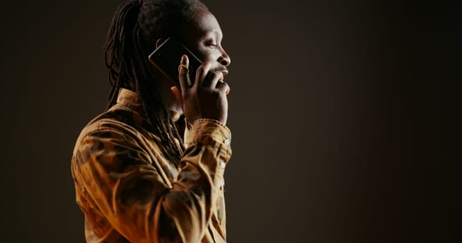 Cool guy with dreads answering phone call in studio, talking to friends or family on camera. African american trendy adult chatting remotely with people on telephone line. Handheld shot.