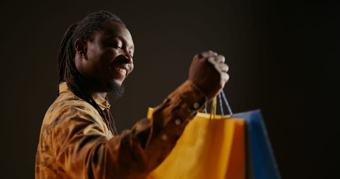 Happy pleased person carrying shopping bags after buying favorite clothes on discount, enjoying sales season at the mall. African american shopaholic guy looks at his new clothing items in bags.