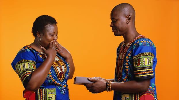 Romantic man offering present with ribbon and bow to his wife, giving giftbox to celebrate her birthday or their anniversary. African american woman receiving sweet wrapped gift from her partner.