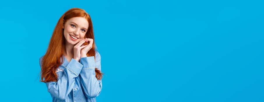 Tenderness, beauty and lifestyle concept. Cute happy charming redhead woman in lovely nightwear, getting ready sleep, wearing pyjama, tilting head and touching face, smiling, blue background.