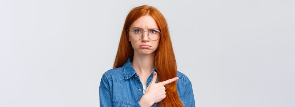 Close-up gloomy and upset, offended sulking girlfriend want something and pointing envy left, indicate disappointing promo, unfair situation, pouting and frowning look displeased, white background.