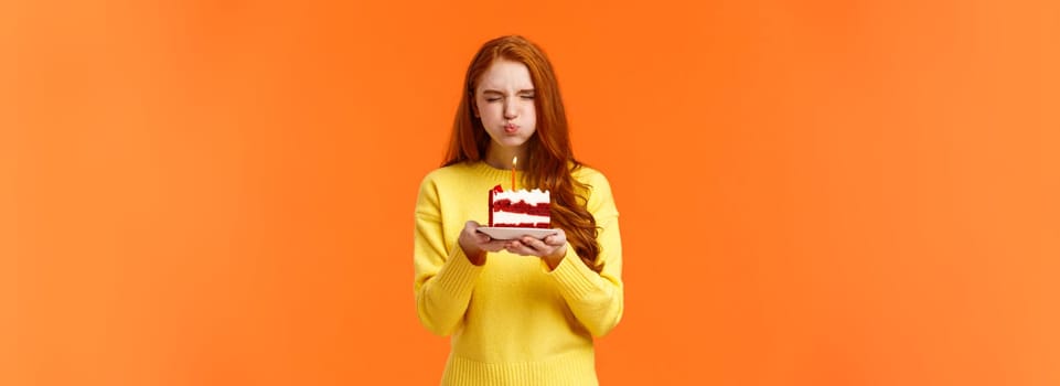 Cute and silly, lovely redhead b-day girl close eyes, holding birthday piece cake, pouting as blow out candle to make wish, want dream come true, close eyes, celebrate with friends, orange background.