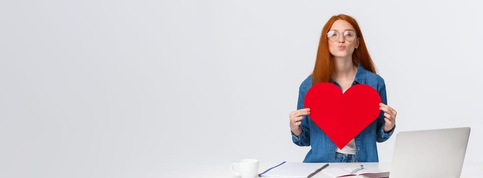 Lovely, romantic cute redhead girl folding lips in mwah, send air kiss and holding big red valentines card, showing her love and passion, congratulate girlfriend with anniversary, stand near laptop.