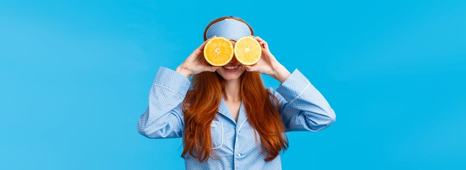 Carefree pretty and cute redhead woman in nightwear, sleep mask, holding two slices of orange over eyes and smiling, fool around in morning, standing blue background joyful.
