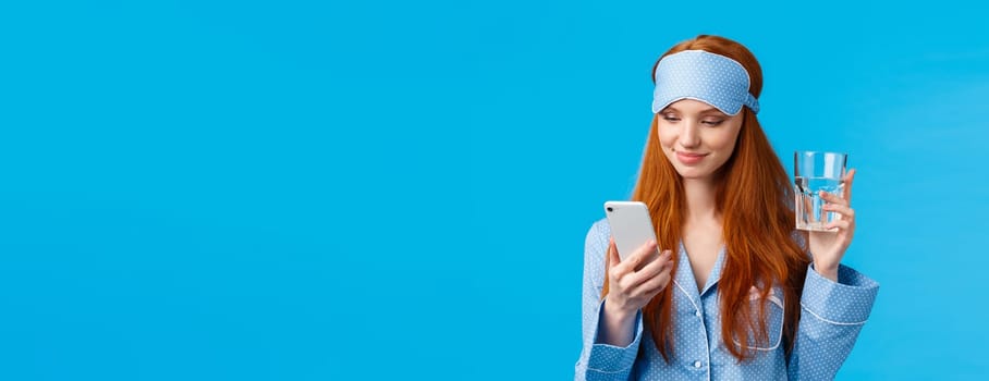 Waist-up portrait joyful redhead european glamour girl in sleep mask and pyjama, holding glass water and checking drinking schedule app, smiling as chatting via smartphone, blue background.