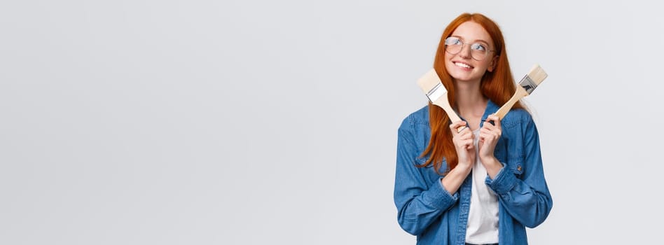 Creativity, imagination and people concept. Good-looking redhead dreamy girl designer, planning to renovate, paint walls in appartment, looking thoughtful and holding paintbrush, white background.