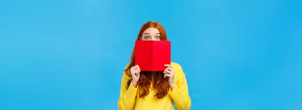 Shy and cute redhead girl like reading books and writing in diarty, hiding face behind red cool notebook, stare curious and surprised with popped eyes, standing blue background modest.