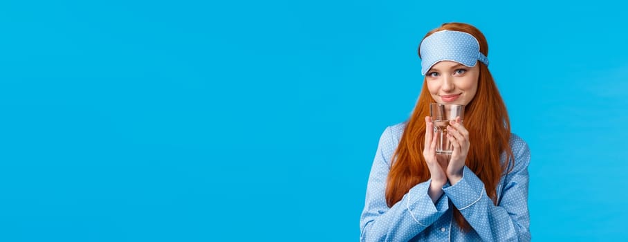 Silly and flirty, feminine redhead woman in cute nightwear and sleep mask, looking coquettish with pleased tender smile, drinking water, start day healthy and active, standing blue background.