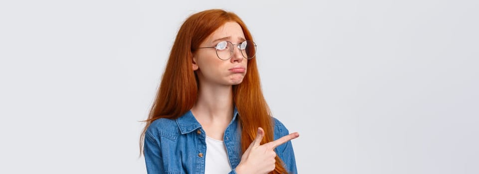 Close-up portrait gloomy and sad pouting cute redhead girl with lovely blue eyes, wear glasses, sulking and sobbing sad, pointing looking upper left corner with regret or jealousy, want something.