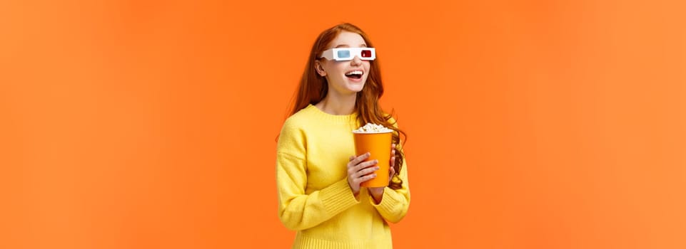 Girl eating popcorn, smiling amused as staring at large screen watching movie at cinema, open mouth thrilled, wear 3d glasses on fantasy film theatre, standing orange background.