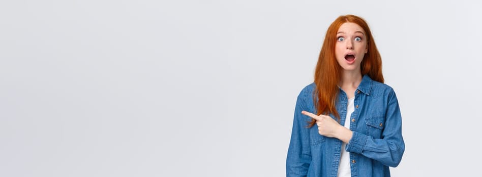 Wow so awesome, check this out. Impressed speechless and fascinated redhead female student drop jaw, looking astounded with popped eyes, pointing finger left, standing white background.