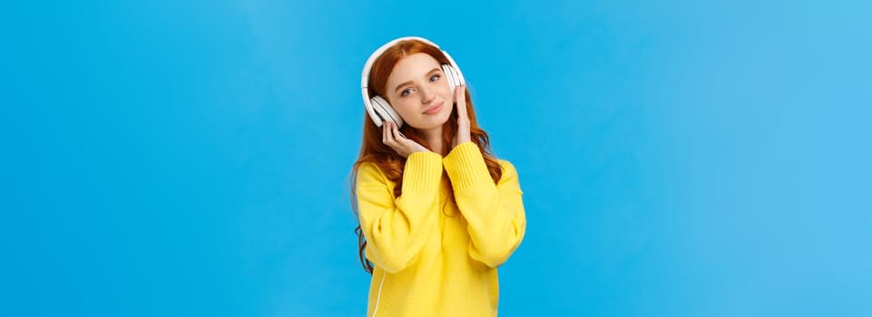 Waist-up shot tender and cute, lovely redhread woman in yellow sweater, tilt head, wear headphones, touching earphones as press to ears, listen music, smiling camera delighted, blue background.