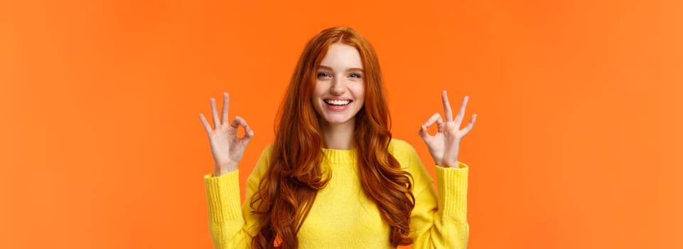 Fashion, shopping, winter holidays concept. Attractive cheerful redhead woman showing okay gestures and smiling, everything alright, approve or like product, recommend online store, orange background.