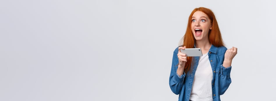 Happy astonished redhead girl cant believe she just won, feeling lucky and satisfied, fist pump, holding mobile horizontally, playing smartphone game, yelling yes, hooray over white background.