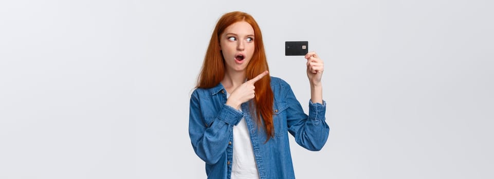 Discounts, shopping and banking concept. Waist-up impressed, thrilled cute redhead girl in denim shirt holding credit card, pointing at it with fascinated impressed eyes, standing white background.